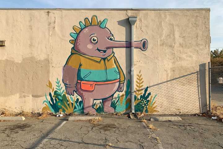 A large painted mural on the stucco wall of a building facing a parking lot, showing a rounded creature with a long snout and a crown of spines standing among painted plants, its snout crossing behind a metal downspout and continuing on the other side