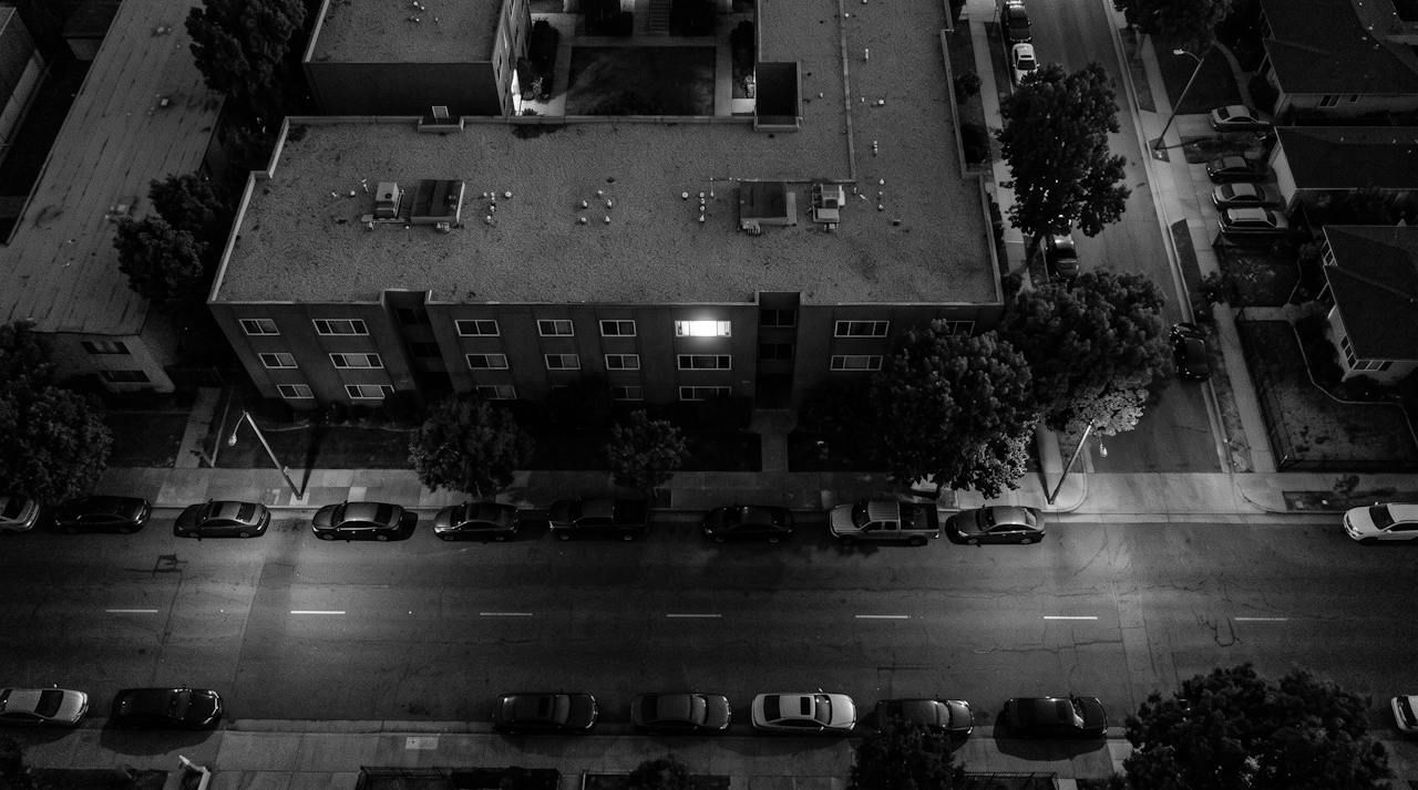 Monochrome overhead surveillance frame of a low apartment block at night, one lit window in a row of dark ones, a street of parked cars
