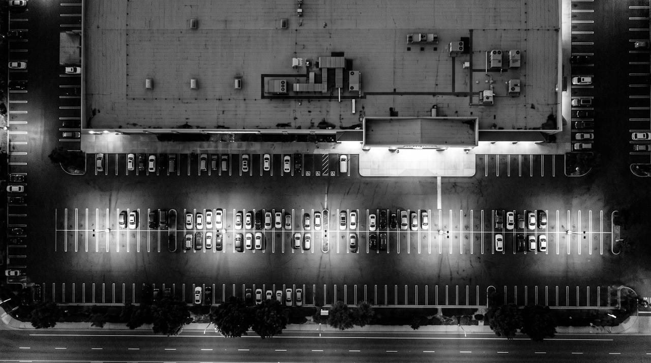 Monochrome overhead surveillance frame of a supermarket parking lot at night, cars in two rows, roof units on the store, a lit entrance canopy
