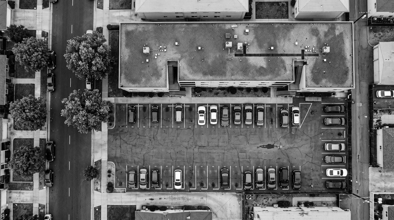 Monochrome overhead surveillance frame of a low apartment block and its parking lot in flat daylight, cars in marked bays along one side, street trees and a residential road