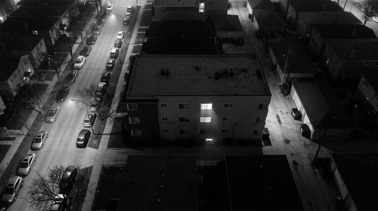Monochrome overhead surveillance frame of a low apartment block at night in haze, two lit windows one above the other, cars parked nose to tail along both kerbs, a service alley behind the building