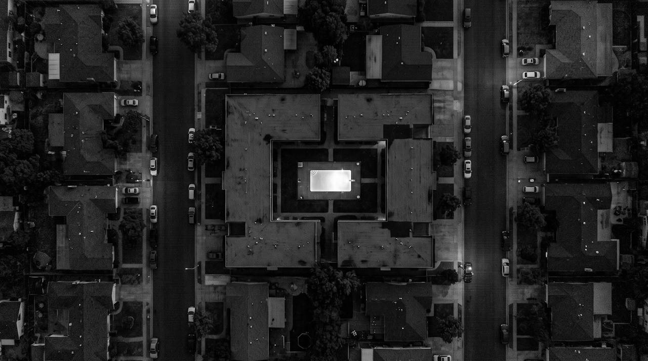 Grainy monochrome overhead image of a residential block at night, a square apartment building closed around an inner courtyard with one small bright rectangle at its centre
