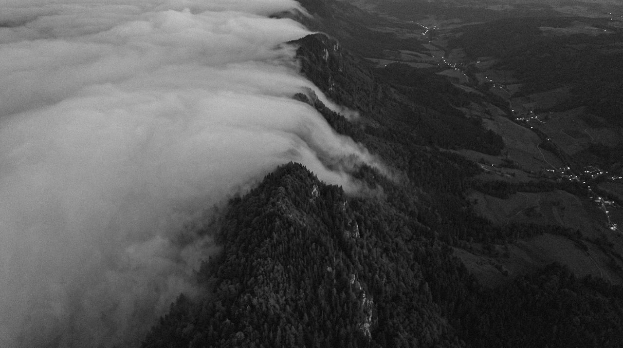 Grainy monochrome overhead image of a forested ridge at dusk, a smooth bank of cloud lying against its far side and spilling over the crest in lobes that fade out partway down the near slope