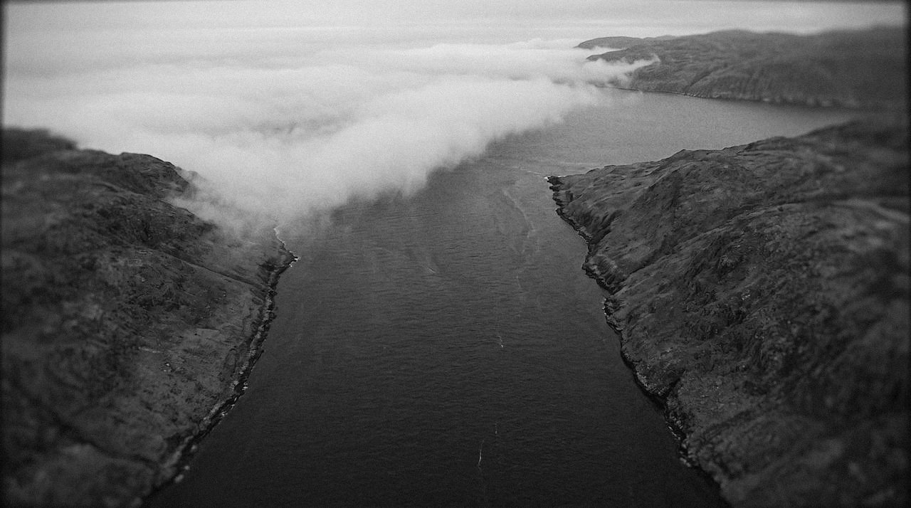 Grainy monochrome overhead image of a narrow strait between hills, a sharp-edged bank of cloud lying across the water and pushing through the gap, the land on either side still clear