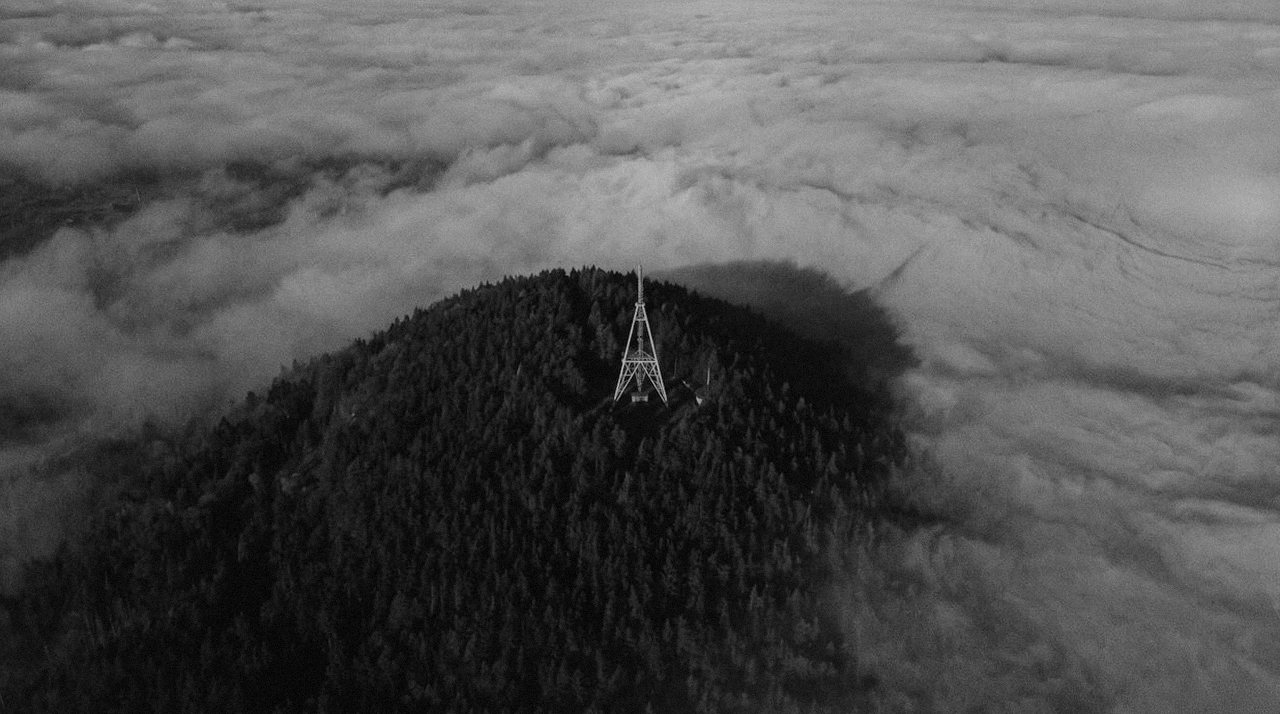 Grainy monochrome image looking down on a wooded summit that rises out of a bank of cloud, a lattice mast standing at the top of it, the hill's shadow lying across the cloud beside it