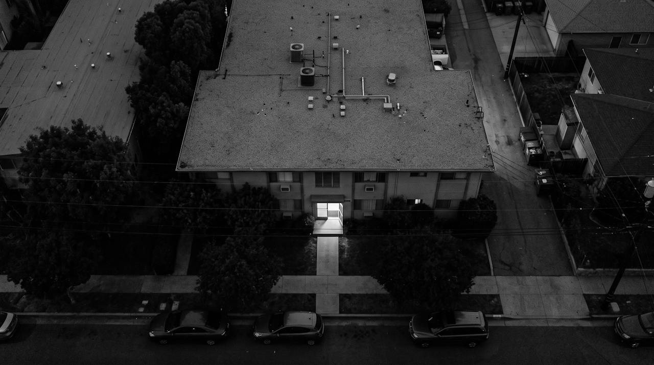 Grainy monochrome image looking almost straight down on the flat roof of a low apartment block at night among street trees, cars parked along the kerb in front, a short walk from the street to the entrance where one small rectangle of light falls on the path
