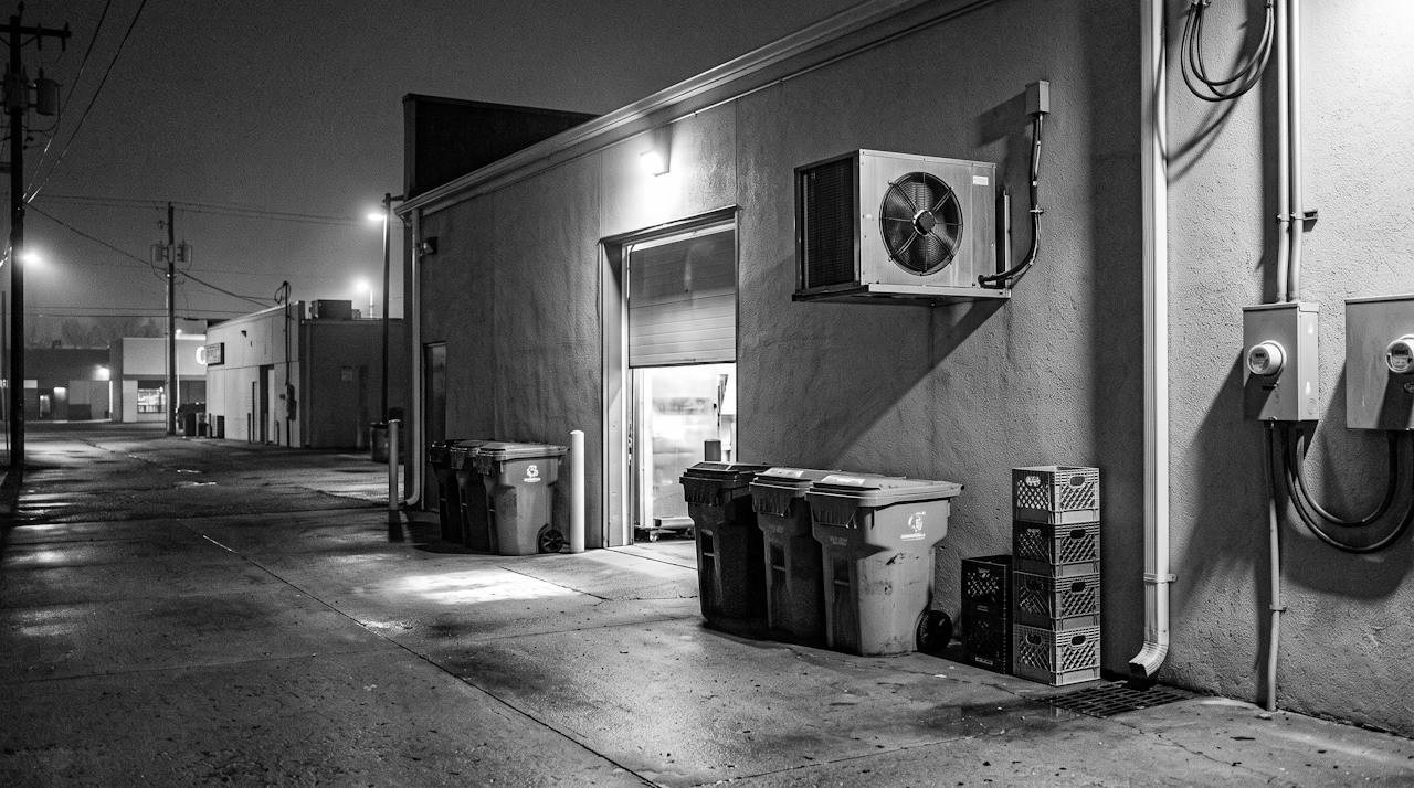 Grainy monochrome long-lens image at night of the back of a restaurant, a loading door propped half open with light spilling out onto wet concrete, a row of bins, a stack of crates, a condenser unit on the wall with its fan grille, sodium haze over the alley